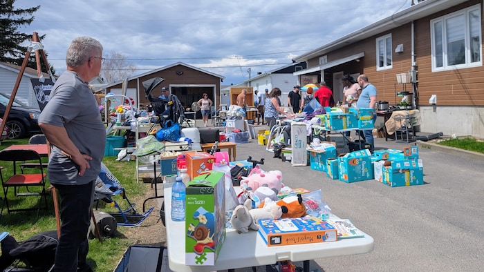 Des gens magasinent à une vente de garage devant une maison, où des tables sont remplies de jouets et d'articles pour bébés.