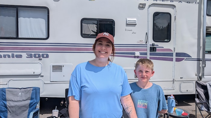 Une femme et un jeune garçon sourient à la caméra. Une roulotte et des chaises de camping sont visibles derrière eux.