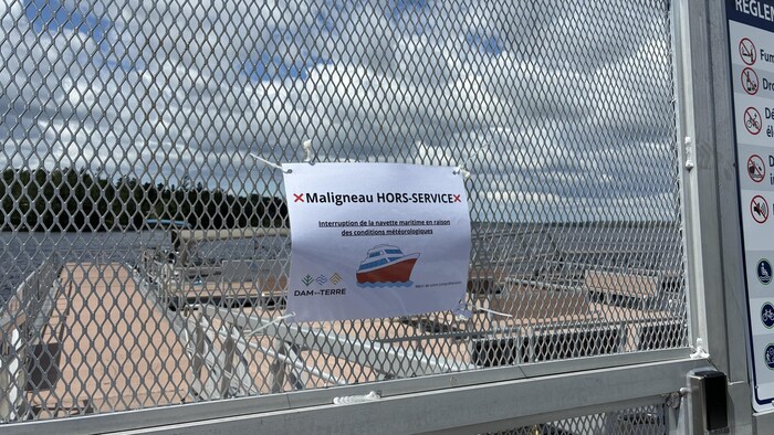 Une affiche blanche fixée à un grillage métallique annonce l'interruption de la navette maritime « Maligneau ». En arrière-plan, à travers la clôture, on aperçoit un quai flottant, un plan d'eau et une rive boisée sous un ciel nuageux.