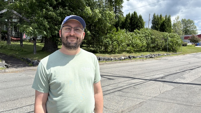 Un homme barbu portant des lunettes, une casquette bleue et un t-shirt vert sourit au premier plan. Il se tient au bord d'une rue asphaltée, devant un arrière-plan composé d'arbres, de buissons et de verdure.