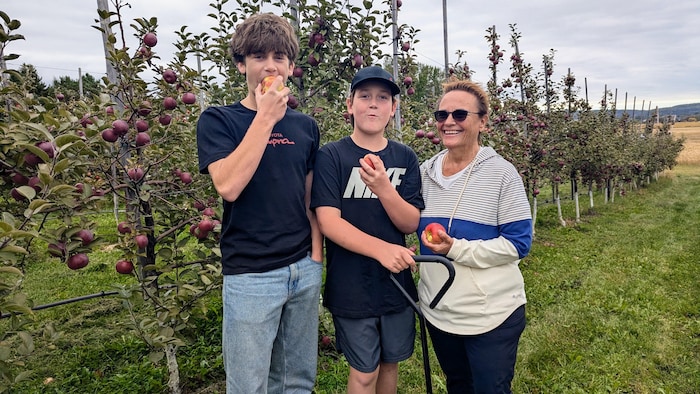 Deux adolescents posent avec leur grand-mère dans une allée de verger, pomme à la main.