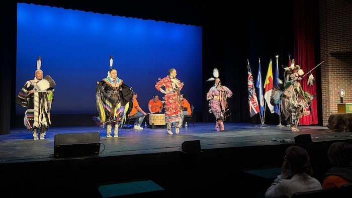 Des danseurs autochtones en habit traditionnel avec des hommes frappant sur un tambour sur une scène.