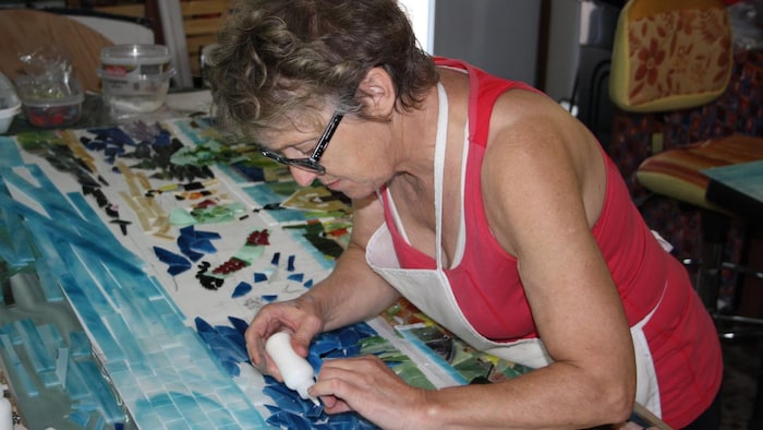 Vue rapprochée de Rose-Marie Gallant, portant des lunettes de lecture et un tablier blanc, penchée sur sa table de travail.