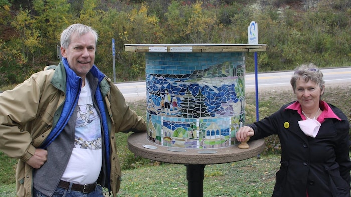 Michel-Caro Tremblay et Rose-Marie Gallant posent de chaque côté d'une structure circulaire en forme de grand touret en bois, ornée d'une fresque minutieuse en mosaïque de verre.