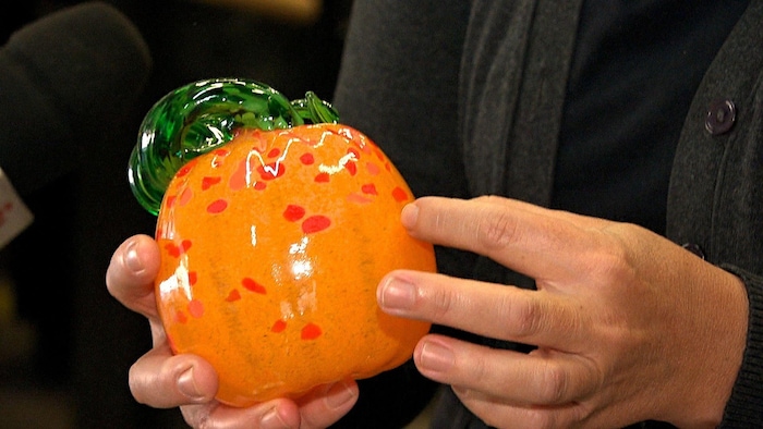 Une artisane tient entre ses mains une citrouille orange et verte en verre soufflé.