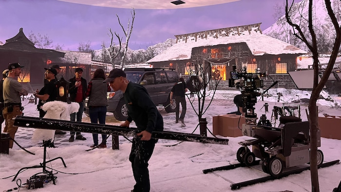 Des hommes travaillent sur un plateau de tournage intérieur. Derrière eux, une image d'un temple japonais en hiver est projetée sur un immense écran.