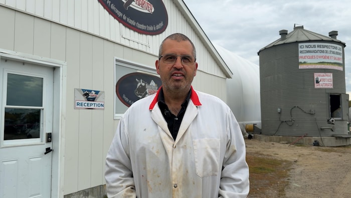 Un homme à l'extérieur face à la caméra devant l'enseigne de la boucherie. Il est en tenu de boucher.