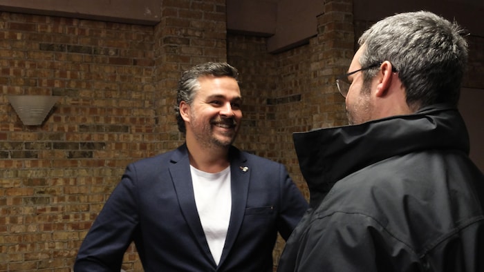 Un homme debout souriant discute avec un autre homme qui est de dos.