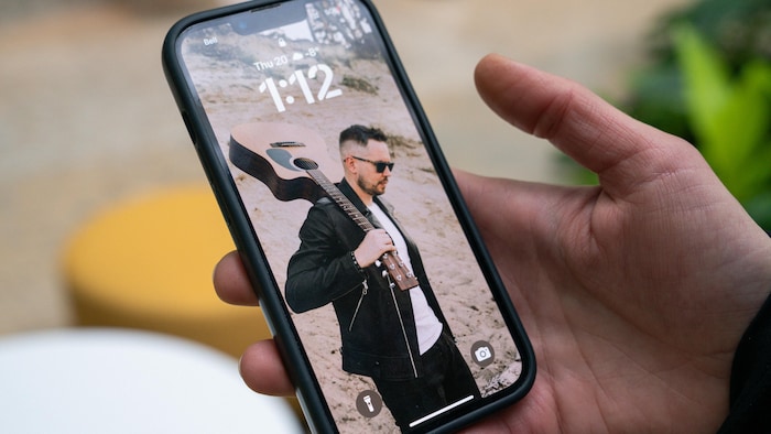 On voit la main de Viktoriia qui tient son téléphone. Sur l'écran, il y a une photo de son mari. Il a les cheveux courts et des lunettes noires et tient une guitare sur son épaule.