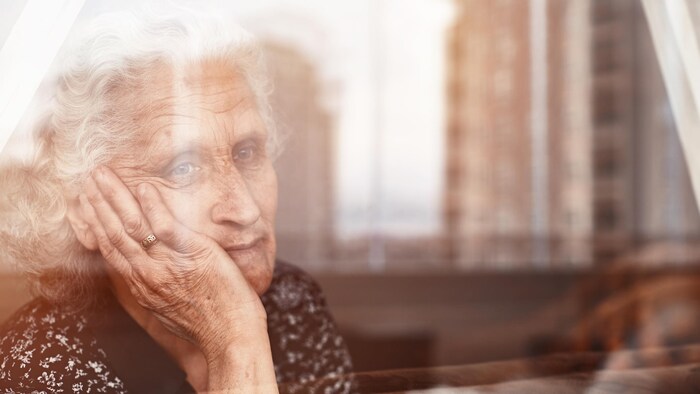 Une dame âgée, la tête tenue dans sa main, regarde par la fenêtre.