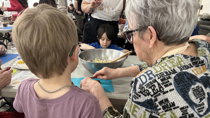 Une dame âgée et un jeune enfant coupent quelque chose à une table. La salle est remplie de gens.