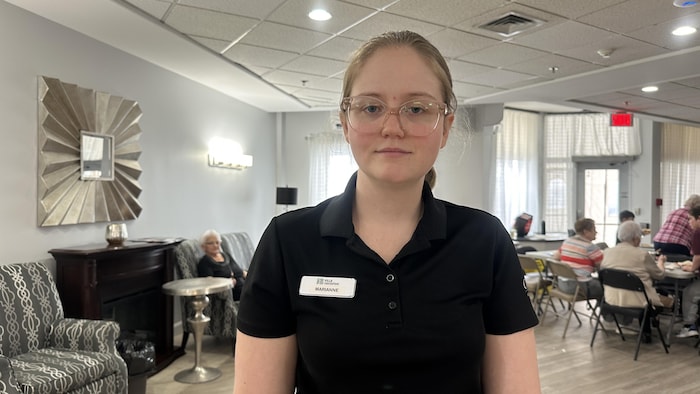 Une jeune femme portant un badge pose pour la caméra dans une grande salle. Des gens sont assis derrière.