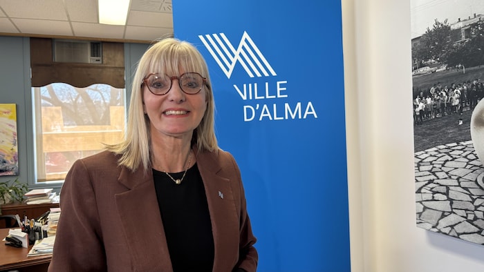 Une femme blonde à lunettes sourit, debout devant une bannière de la ville d'Alma.