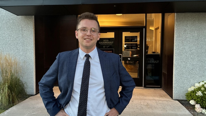 Un homme en complet et cravate sourit à la caméra devant la porte d'un bâtiment commercial.