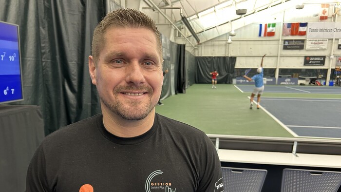 Vincent Milot se tient debout à quelques pas d'un terrain de tennis pendant que des joueurs disputent une partie.