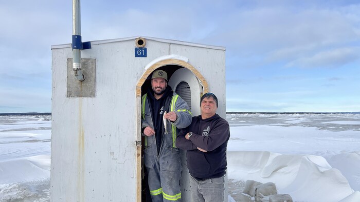 Deux personnes devant une cabane sur la banquise. L'un d'eux tient un petit poisson.