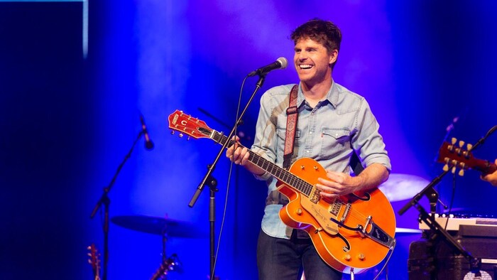 Vincent Vallières, guitare à la main qui arbore un grand sourire sur la scène de l'Aphithéâtre Cogeco.
