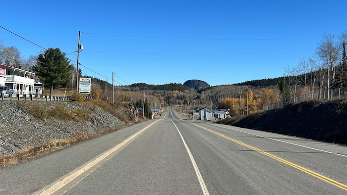 La route et quelques bâtiments d'un village, avec une montagne en forme de grosse bosse au loin.