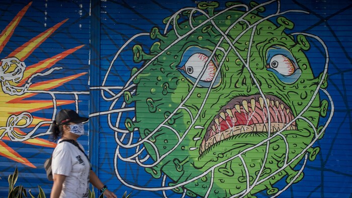 Une femme masquée passe devant une murale d'un virus qui a l'air menaçant.