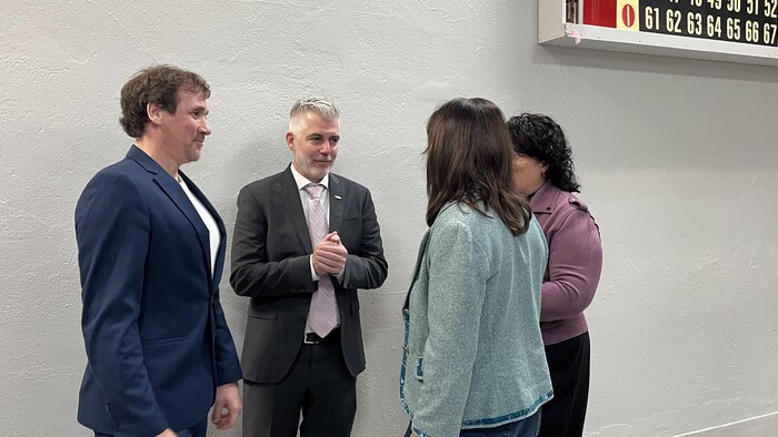 Quatre personnes, deux hommes et deux femmes en tenue professionnelle, discutent debout près d'un mur gris. À droite, un tableau de bingo électronique est visible. L'ambiance suggère une conversation informelle lors d'un événement officiel ou communautaire.