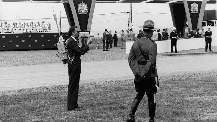 Un caméraman de Radio-Canada, portant son équipement sur son dos et des membres de la Gendarmerie royale du Canada sur le site de la cérémonie d'inauguration de la Voie maritime du Saint-Laurent.