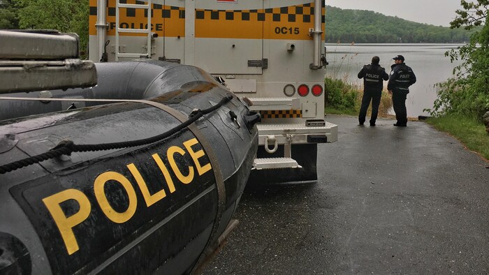 Un camion de la Sûreté du Québec transportant derrière lui un bateau Zodiac avec l'inscription Police est stationné au bord d'un lac. 