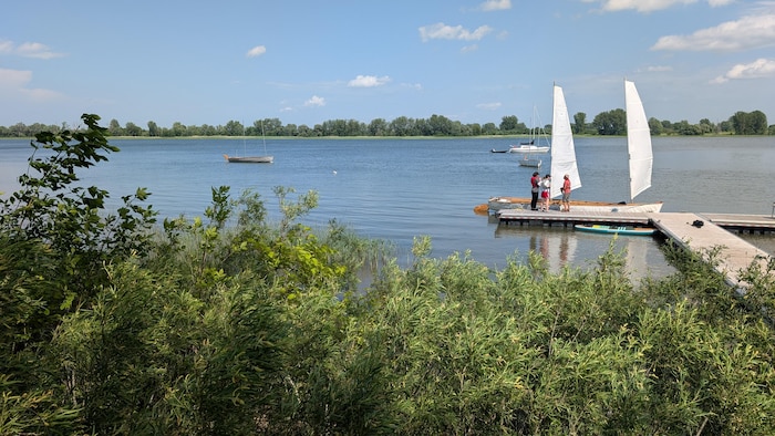 Au bord du fleuve Saint-Laurent, trois personnes sont debout sur un quai devant un petit voilier à deux mâts.