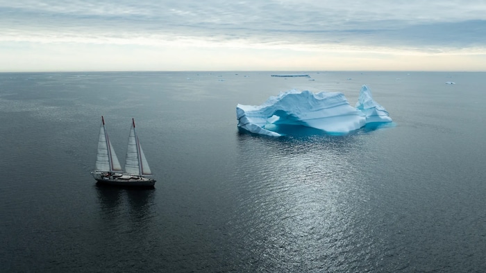 Vue aérienne d'un voilier sur une grande étendue d'eau où flotte un iceberg.
