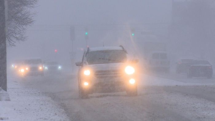 Une voiture aux phares allumées roule sur une route enneigée alors qu'il neige tout autour.