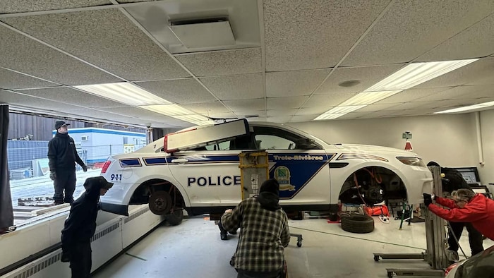 Cinq hommes s’affairent autour d’un véhicule de la police de Trois-Rivières, dont les roues sont enlevées, à l’intérieur d’un local.