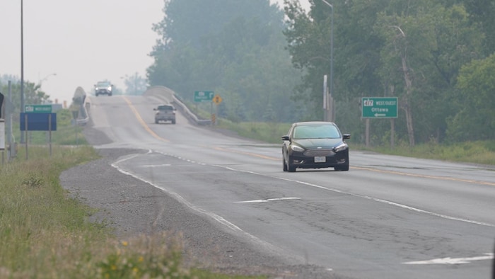 Une petite voiture a la sortie de l'autoroute 417 durant la journée à l'été et un viaduc en arrière-plan avec deux camions.