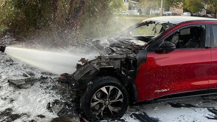 Une voiture calcinée se fait arroser par un boyau de pompier.