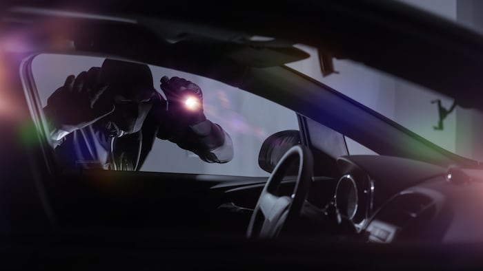 Un individu cagoulé dirige la lueur d'une lampe de poche vers l'intérieur d'une automobile stationnée.