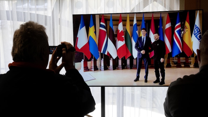 Un vidéaste filme un écran de télévision sur lequel apparaissent Trudeau et Zelensky, qui se serrent la main.