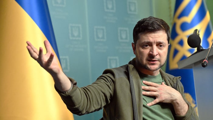 Volodymyr Zelensky, en tenue kaki, devant des drapeaux ukrainiens.