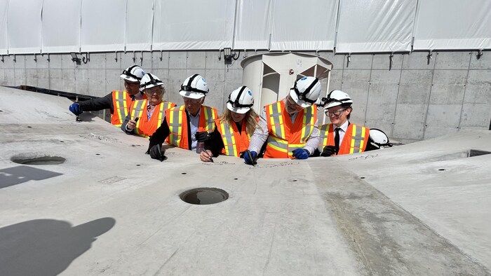 Séance de signature d’un voussoir par six personnes, dont les ministres Chantal Rouleau et Benoit Charettte, qui portent des casques protecteurs et des vestes de sécurité pendant une visite de chantier.