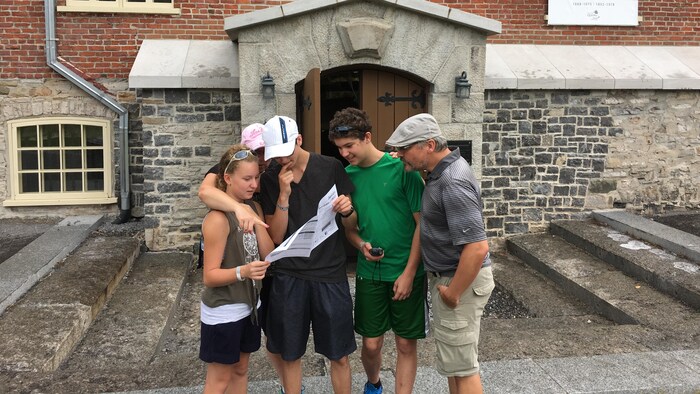 Une famille analyse un document, appareil GPS en main, devant un vieil immeuble en pierres et en briques.