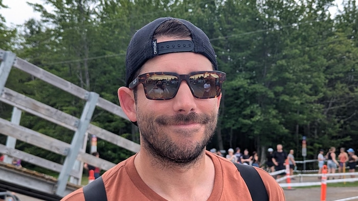 Un homme qui porte des lunettes fumées dans lesquelles se reflètent, dans la lentille de gauche, la journaliste qui le prend en photo, et dans la lentille de droite, un caméraman qui tient son trépied.