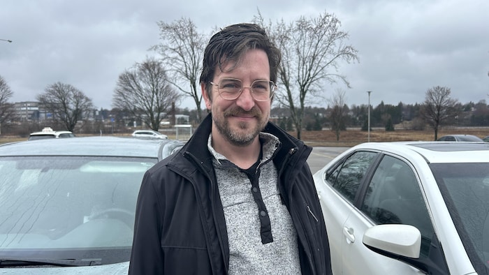 Un homme portant des lunettes sourit en posant pour une photo dans un stationnement, entre deux voitures.
