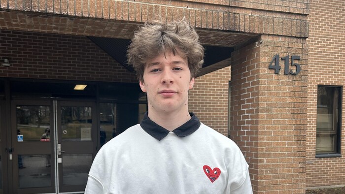 Un jeune homme portant un chandail blanc décoré d'un coeur rouge est debout devant un édifice en briques.