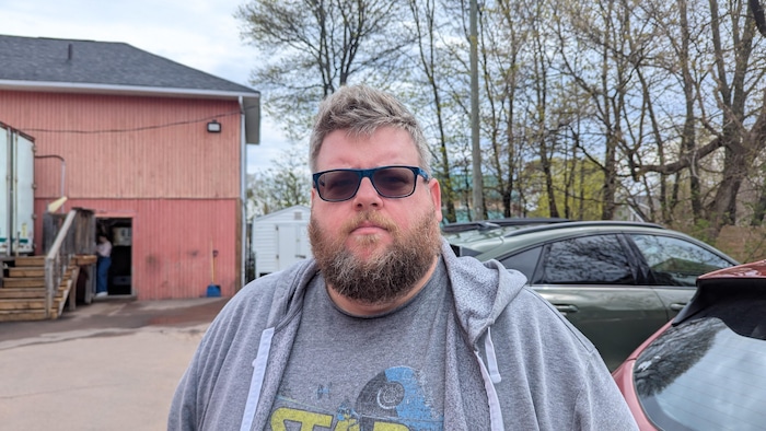 Un homme portant une barbe et des lunettes fumées, avec un bâtiment et des autos stationnées derrière.