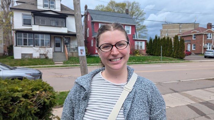 Une femme portant des lunettes est debout sur le trottoir d'un quartier résidentiel et fait un grand sourire.