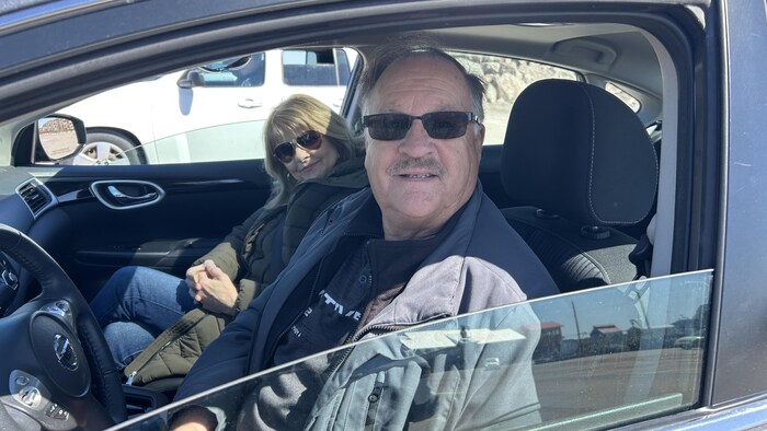 Un homme portant des verres fumés et une moustache est assis au volant de son véhicule, la vitre baissée. Il y a une femme dans le siège passager.