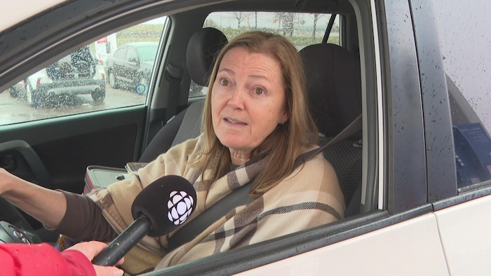 Une femme assise au volant de son véhicule aux vitres maculées de gouttes de pluie. Quelqu'un pointe un micro de Radio-Canada vers elle.