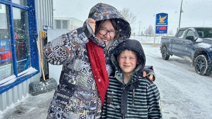 Devant une station-service pendant une tempête de neige, une femme tient le capuchon de son manteau d'hiver avec sa main droite, et elle a le bras gauche autour des épaules de son petit-fils.