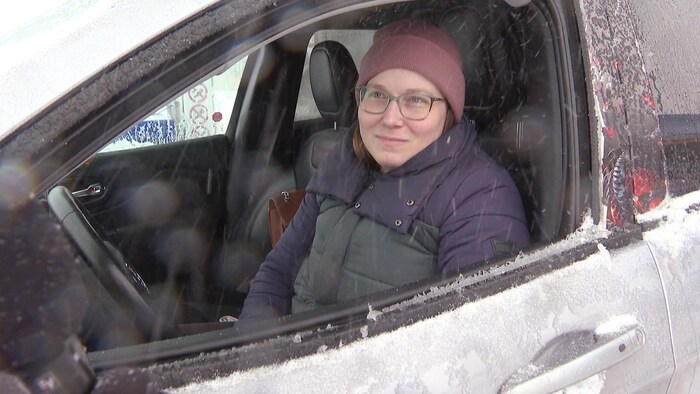 Une femme portant une tuque et un manteau d'hiver est assise dans le siège conducteur de son automobile, la fenêtre grande ouverte pendant une tempête de neige.