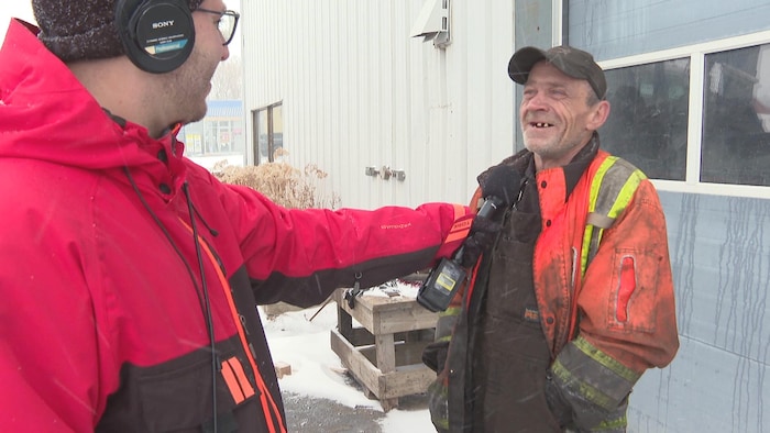 Un homme en salopette et manteau de travail est dehors pendant une tempête de neige et jase avec le journaliste.