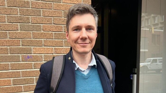 Un homme pose pour une photo sur le trottoir, debout devant un mur de briques.