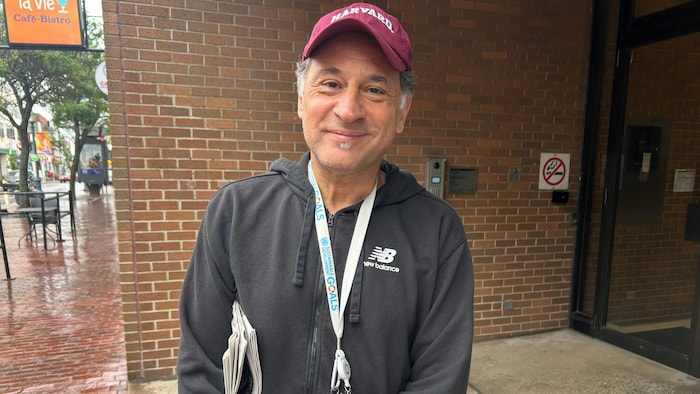 Un homme avec un chandail à capuchon New Balance, une casquette de l'université Harvard et un journal sous le bras pose pour une photo dehors dans l'entrée d'un immeuble à bureaux. 