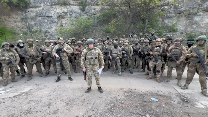 Un homme qui porte un uniforme militaire et qui tient une arme automatique est debout devant des dizaines d'hommes en treillis militaires.